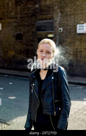 Frau, die Rauch ausatmet, während sie im Sonnenlicht gegen eine Ziegelwand steht und eine schwarze Lederjacke trägt. Bola02840 Copyright: XConnectxImagesx Stockfoto