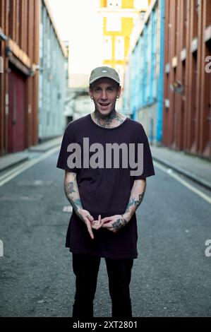 Lächelnder tätowierter Mann in einem dunklen T-Shirt und Mütze, der in einer städtischen Gasse steht. Bola02834 Copyright: XConnectxImagesx Stockfoto