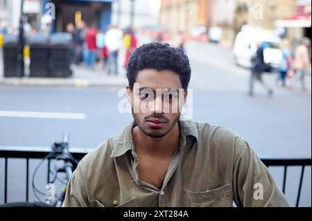 Ein junger Mann mit lockigen Haaren schaut ruhig in die Kamera, mit einer verschwommenen Straßenszene im Hintergrund. Bola02865 Copyright: XConnectxImagesx Stockfoto