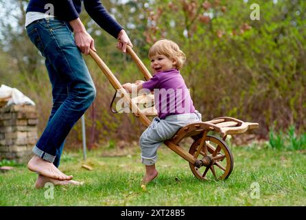 Ein Kleinkind fährt im Freien in einem Holzwagen, der von einem Erwachsenen an einem sonnigen Tag gezogen wird. Bola02899 Copyright: XConnectxImagesx Stockfoto