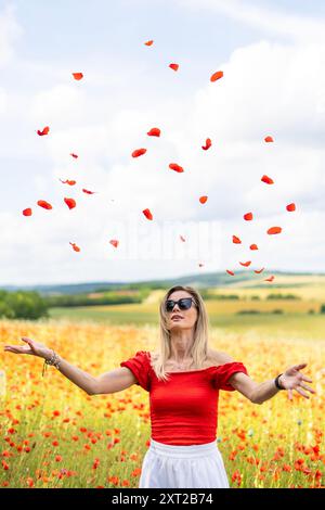 Blonde junge kaukasische Frau in rot-weißem Kleid und Sonnenbrille streut rote Blütenblätter in der Luft auf einem Mohnfeld Stockfoto