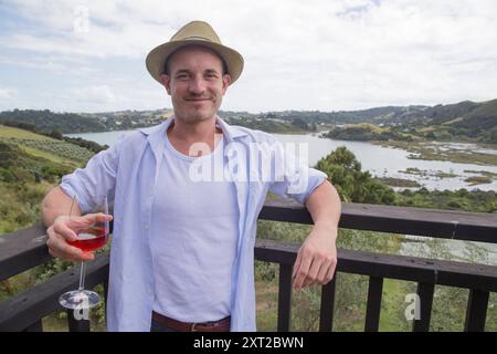 Lächelnder junger Mann mit Fedora-Hut, der draußen ein Glas Wein hält und im Hintergrund einen malerischen Blick auf den See hat, Auckland, Neuseeland evpe01265 Polizist Stockfoto