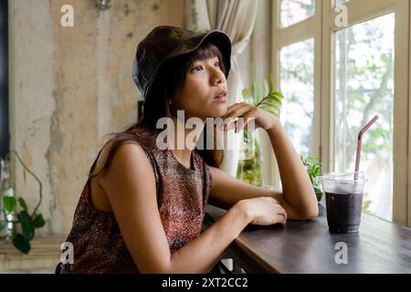 Junge asiatische Frau mit Hut, die aus dem Fenster eines Cafés warmes Ambiente blickt Stockfoto