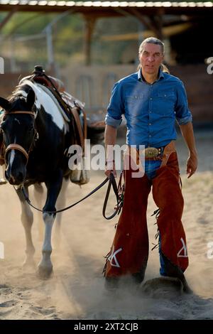 Ein selbstbewusster Mann in Cowboykleidung geht neben einem Sattelpferd in einer staubigen Arena. Bola03080 Copyright: XConnectxImagesx DATENSATZDATUM NICHT ANGEGEBEN Stockfoto