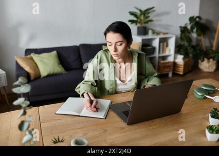 Eine Frau sitzt zu Hause an einem Schreibtisch und schreibt in ein Notebook, während ihr Laptop vor ihr offen ist. Stockfoto
