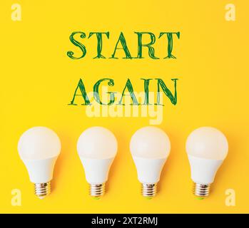 Vier weiße Glühlampen in einer Reihe auf gelbem Hintergrund. Das Wort „Start Again“ wird in grünen Buchstaben über den Lampen geschrieben, wodurch ein Gefühl von entsteht Stockfoto