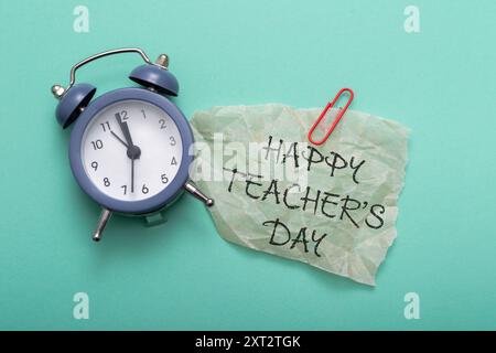 Eine Uhr ist auf grünem Hintergrund mit einem Blatt Papier, auf dem steht "Happy Teacher's Day". Die Uhr ist auf 10:00 Uhr eingestellt Stockfoto