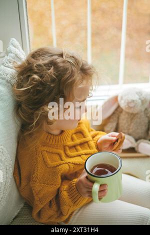 Das kleine süße, lockige Mädchen im gestrickten Pullover trinkt Tee und isst Kekse vor dem Fenster. Gemütliches Zuhause. Warmer Herbst. Stimmung. Stockfoto