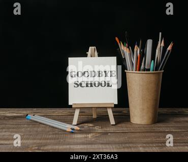 Ein Schild, auf dem die Schule auf Wiedersehen steht, steht auf einem Holztisch. Neben dem Schild sind Bleistifte und eine Tasse Bleistifte Stockfoto