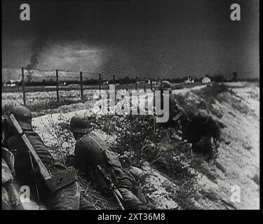 Vier männliche Soldaten der deutschen Armee liegen niedrig auf einem Hang unter einem Zaun, während Rauchsäulen aus einer Baumreihe in der Entfernung hinter mehreren Häusern aufsteigen, 1939. Aus „Time to Remember – The Reluctant Warriors“, 1939 (Reel 3); Dokumentarfilm über Ereignisse von 1939 – Vorbereitungen für den Krieg und dann Feindseligkeiten brechen aus. Stockfoto