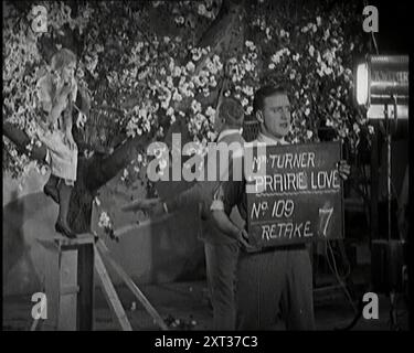 Weibliche Schauspielerin sitzt in einem Blossom Tree, während ein männlicher Zivilist mit ihr spricht. Ein Filmtechniker hält ein Clapper Board mit der Aufschrift „Mr. Turner Prairie Love No. 109 Rake 7“, 1920er Elstree, im Großen und Ganzen das Hollywood Großbritanniens in den Zwanzigern, aber ohne Palmen und Sonne. Aber auf den Bühnen mit Zinndach konnte man es zu jeder Jahreszeit schaffen. Aus „Time to Remember – Come the Dawn“, 1925 (Rolle 1); werfen Sie einen Blick auf die britische Filmindustrie der 1920er Jahre – Clips aus großen Stummfilmen und Wochenschauen. Stockfoto