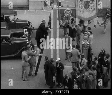 Bud Flanagan mit einer Bratpfanne, die eine Leiter klettert, die vor Krönungsdekorationen in der Oxford Street steht, während Zuschauer zuschauen, und Chesney Allen klettert hinter ihm mit einem dritten Mann, 1937. "Letzte Berührung der Dekorationen [in London]. Aber wer steckt hier ihr Ruder - oder besser gesagt ihre Leiter - hinein? Flanagan und Allan, nachdem sie entdeckt haben, dass einige der Posts Fackeln unterstützen, beschließen sie, Treibstoffrechnungen zu sparen, indem sie in der Oxford Street kochen. Aus „Time to Remember – Sense of Values“, 1937 (Reel 2); Dokumentarfilm über die Ereignisse von 1937, Krieg in der Fernen EA Stockfoto