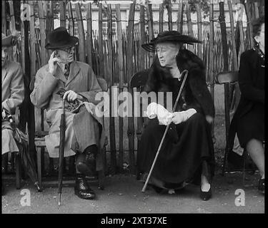 Eine ältere Frau mit breitem Hut, einem Fellstohlen und einem Wanderstock, der auf einer Bank vor einem Zaun sitzt und mit einem älteren Mann spricht, der einen langen Mantel trägt und einen Regenschirm hält, der neben ihr sitzt, 1938. Aus „Time to Remember – Wind Up Week“, 1938 (Reel 1); Dokumentarfilm über 1938 – die Menschen werden sich der wachsenden Bedrohung des Krieges bewusst. Stockfoto