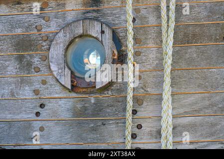 Nahaufnahme eines verwitterten Holzschneiders mit rundem Bullauge und geflochtenen Seilen. Stockfoto