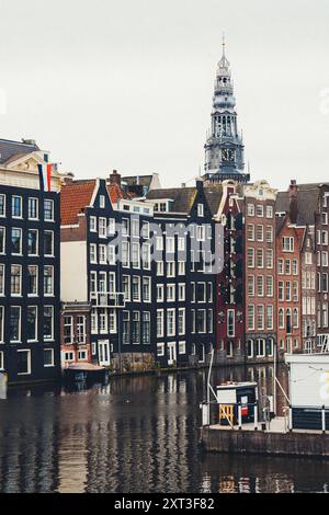 Ein ruhiger Blick auf den Amsterdamer Kanal, gesäumt von markanten, schmalen Gebäuden und dem kunstvoll verzierten Turm einer alten Kirche, der sich darüber erhebt. Die Szene erfasst den Stockfoto