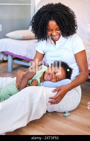 Eine fröhliche afroamerikanische Mutter wird lachend dargestellt, während sie mit ihrer kleinen Tochter spielt, die auf einem Kissen liegt und beide einen verspielten Moment genießen Stockfoto