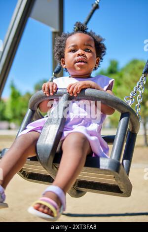 Ein entzückendes afroamerikanisches Kleinkind, gekleidet in einem hellen lila Kleid, sitzt an einem hellen sonnigen Tag auf einer Schaukel in einem Park Sie sieht zufrieden und curi aus Stockfoto