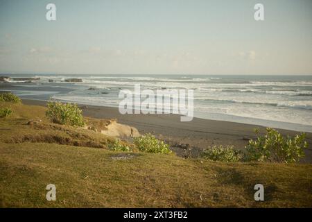 Blick auf das tropische Meer mit Windwellen von oben auf einem grasbewachsenen Hügel am Rand eines schwarzen Sandstrandes Stockfoto