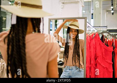 Eine junge Frau lächelt sich im Spiegel an, während sie in einem Boutique-Bekleidungsgeschäft einen Strohhut ansetzt und mit freudigem Ausdruck in die Kamera schaut. Stockfoto