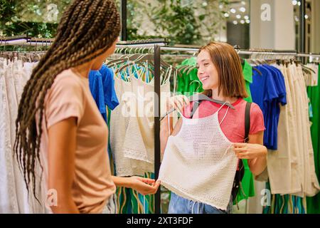 Zwei junge multiethnische Frauen, eine mit geflochtenen Haaren und die andere mit kurzen Haaren, sehen sich an und lächeln, während sie eine Häkelspitze hineinhalten Stockfoto
