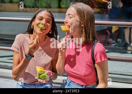 Zwei multirassische Freundinnen teilen sich einen freudigen Moment beim Essen frischer Obsttassen im Freien. Sie lächeln und zeigen eine starke Freundschaft und eine lässige Stockfoto