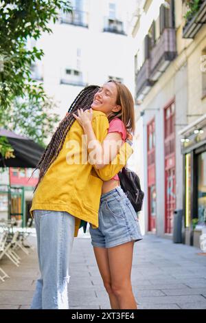 Seitenansicht von zwei Frauen, die glücklich auf einem sonnendurchfluteten Bürgersteig umarmen, beide Ausdruck von Freude und Zuneigung. Die Frau in einer gelben Jacke schaut weg, di Stockfoto