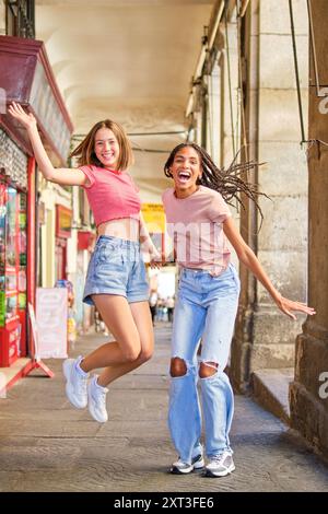 Zwei glückliche junge Frauen, die energisch auf einer belebten Stadtstraße springen und lachen, feiern gemeinsam einen freudigen Moment. Beide schauen sich das Kommen an Stockfoto