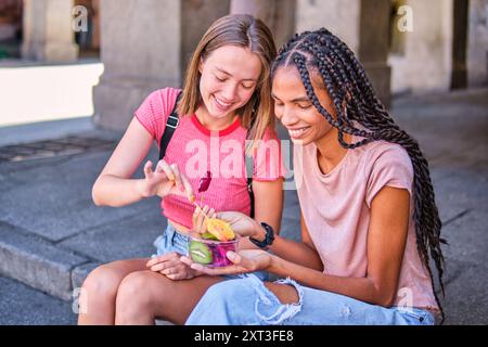 Zwei Freundinnen teilen sich eine bunte Schüssel mit frischem Obst, während sie draußen sitzen. Eine Freundin schaut auf die Schüssel, während sie ein Stück aussucht, und das Stockfoto