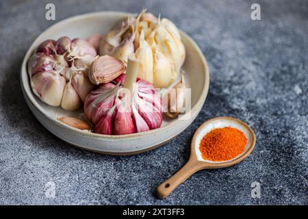 Eine Nahaufnahme frischer Knoblauchzwiebeln und Nelken in einer Keramikschale neben einem Holzlöffel lebendiger Gewürze, vor einem strukturierten grauen Backgr Stockfoto