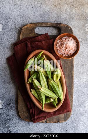 Frische Okra-Bamia-Schoten werden in einer Terrakotta-Schüssel auf einem hölzernen Schneidebrett auf einem strukturierten grauen Hintergrund präsentiert, der den rustikalen Charme verkörpert Stockfoto