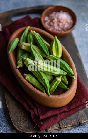 Frische, ungekochte Okra-Bamia-Schoten in einer traditionellen Holzschale, kombiniert mit einer Tonuntertasse aus rosa Himalaya-Salz, auf einer rustikalen Kücheneinrichtung Stockfoto