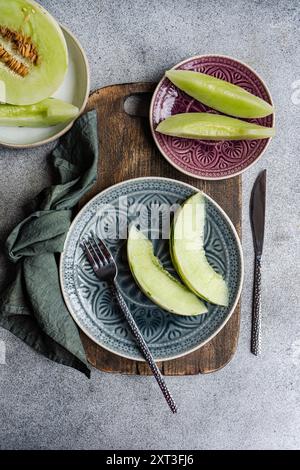Blick von oben auf frisch geschnittene Bio-Melonen, die elegant auf dekorativen Tellern angeordnet sind, begleitet von einer dunklen Metallgabel und einem rustikalen Holzbrett, Re Stockfoto