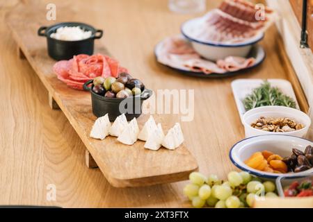 Reichhaltige Auswahl an Gourmet-Vorspeisen, die ordentlich auf einem langen Holzbrett angeordnet sind. Der Aufstrich umfasst geschnittenes Wurstfleisch, Brie-Käse-Wedges, eine Schüssel mi Stockfoto