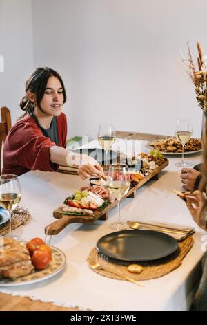 Eine Frau greift auf einer gemeinsamen Charcuterie-Brett auf einer Party zu Hause nach Essen, umgeben von Freunden und Gläsern Wein. Der Tisch ist vielfältig Stockfoto