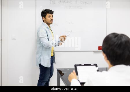 Ein männlicher Lehrer führt eine Lektion durch, während er mit seinen Schülern in einem hellen Klassenzimmer interagiert und seine Punkte mithilfe eines Whiteboards veranschaulicht Stockfoto