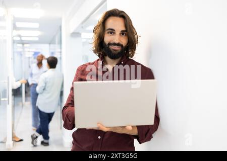 Ein lächelnder männlicher Erzieher mit Bart hält einen Laptop in einem belebten Akademiekorridor, während Kollegen im Hintergrund interagieren Stockfoto
