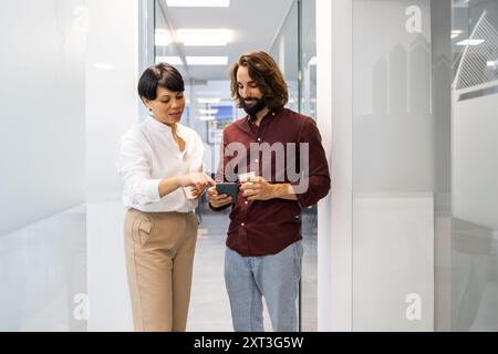 Zwei Profis, ein Mann und eine Frau, nehmen an einer gemeinsamen Diskussion Teil, während sie ein Smartphone in einem hellen, modernen Flur betrachten Stockfoto