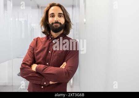 Ein selbstbewusster und naher männlicher Lehrer steht in einem hellen, modernen Flur einer englischen Akademie, mit einem einladenden Auftreten und überkreuzten Armen Stockfoto