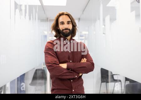 Ein selbstbewusster und naher männlicher Lehrer steht in einem hellen, modernen Flur einer englischen Akademie, mit einem einladenden Auftreten und überkreuzten Armen Stockfoto