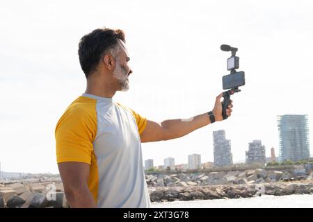 Mann in gelbem T-Shirt, der ein Gimbal mit Smartphone benutzt, um ein Video am Meer aufzunehmen, auf den Bildschirm zu schauen, mit urbaner Skyline im Hintergrund. Stockfoto