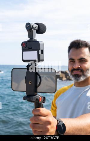 Indischer Mann, der Smartphone und Kamerabildstabilisator benutzt, um neben dem Meer Videos aufzunehmen und auf den Bildschirm zu schauen. Stockfoto