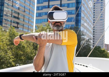 Indischer Mann in Sportkleidung verwendet ein VR-Headset und eine Smartwatch für ein interaktives Workout auf einer sonnigen Stadtbrücke Stockfoto