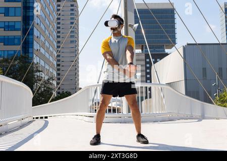 Ein Indianer wird mit einem Virtual-Reality-Headset verwickelt, während er auf einer sonnendurchfluteten Brücke in einer Stadt steht und von der Kamera weg zeigt. Stockfoto