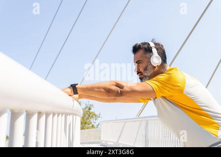 Ein Indianer in einem gelben T-Shirt streckt sich auf einer modernen Brücke, trägt Kopfhörer und eine Smartwatch. Er blickt von der Kamera weg, konzentriert sich auf seine Exkursion Stockfoto