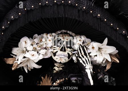 Catrina ist mit einer weißen Blumenmaske und einem schwarzen Outfit verziert und repräsentiert die lebhafte Feierlichkeit zum Tag der Toten in Mexiko Stockfoto