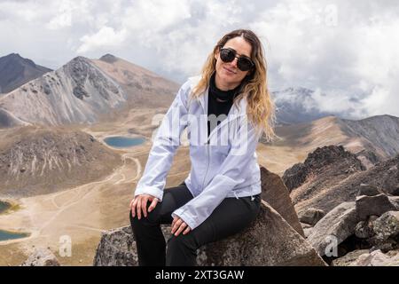 Eine kaukasische Frau mit Sonnenbrille ruht auf einem felsigen Gipfel und blickt in die Kamera, mit einem atemberaubenden Blick auf einen ruhenden Vulkan und das umliegende Land Stockfoto