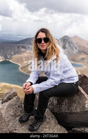 Eine kaukasische Frau mit Sonnenbrille ruht auf einem felsigen Gipfel und blickt in die Kamera, mit einem atemberaubenden Blick auf einen ruhenden Vulkan und das umliegende Land Stockfoto