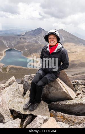 Ein glücklicher Wanderer, lateinamerikanisches Weibchen, sitzt auf einem Felsvorsprung mit Blick auf einen atemberaubenden vulkanischen Kratersee in Mexiko. Sie schaut direkt auf den Camer Stockfoto