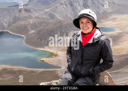 Ein glücklicher Wanderer, lateinamerikanisches Weibchen, sitzt auf einem Felsvorsprung mit Blick auf einen atemberaubenden vulkanischen Kratersee in Mexiko. Sie schaut direkt auf den Camer Stockfoto