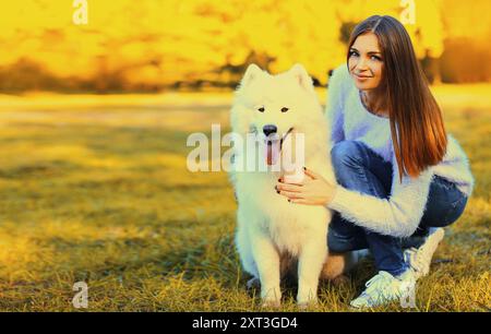 Glückliche lächelnde Frau und weißer Samoidenhund, der auf dem Gras im sonnigen Herbstpark liegt Stockfoto